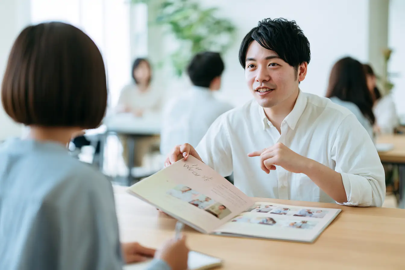 会社説明会のあとの座談会で、若手社員が採用パンフレットを開いて学生に仕事の流れを説明し、学生が熱心に聞いている様子