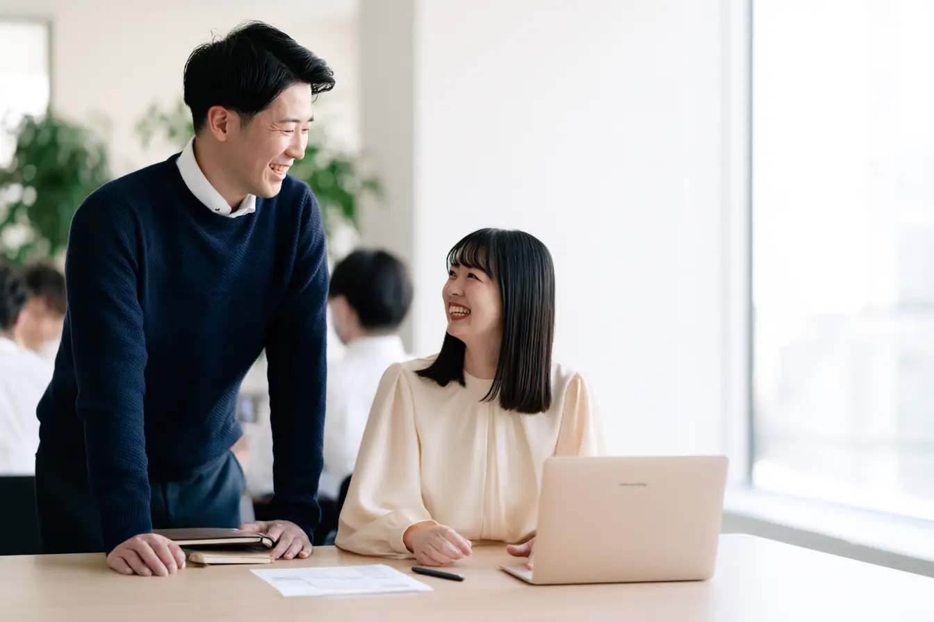 入社して数か月の若手社員が、自分のデスクで先輩と仕事の進め方を相談しながら、自然に笑い合っている様子